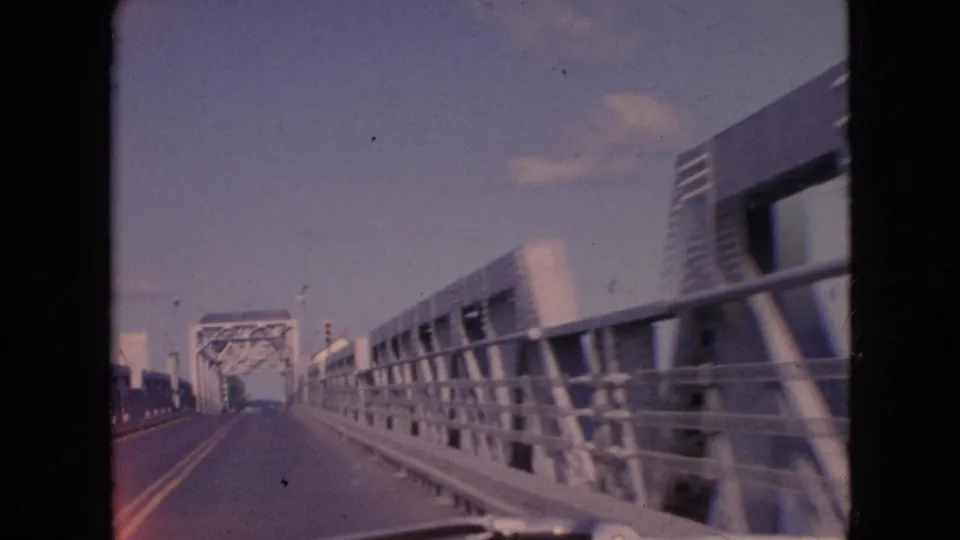 First-person bridge-driving view from the Niagara Falls, New York home-movie clip, showing a paved road, metal railings, and a steel truss span in the distance.