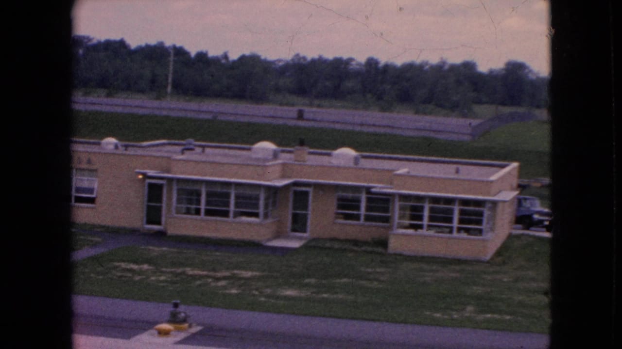 1964 Niagara Falls home-movie frame showing a low beige institutional-looking building with broad windows, grass, a road, and trees in the background.
