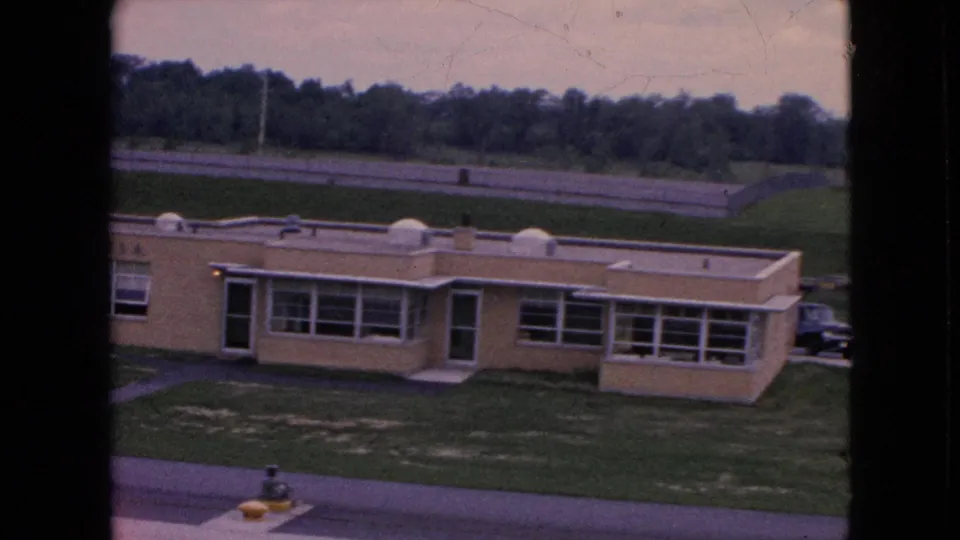 1964 Niagara Falls home-movie frame showing a low beige institutional-looking building with broad windows, grass, a road, and trees in the background.