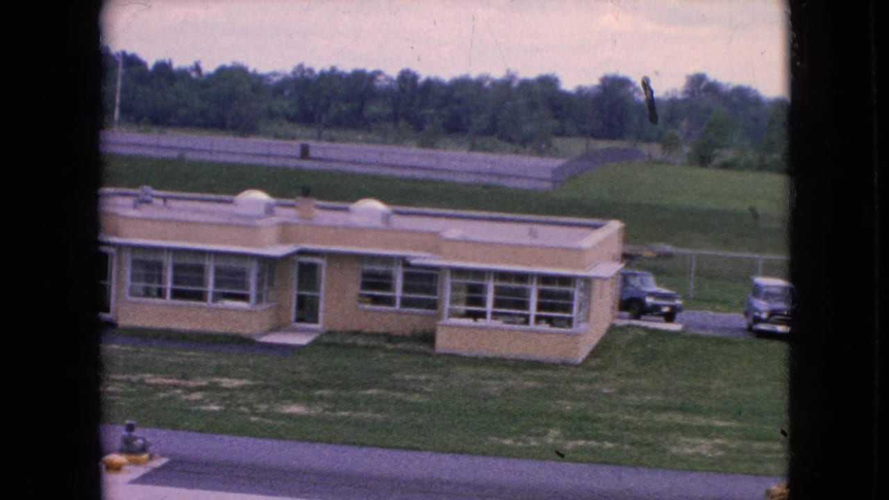 1964 Niagara Falls frame with a beige flat-roofed institutional building, large window bays, a sloped lawn, roadway, distant trees, and parked vintage cars nearby.