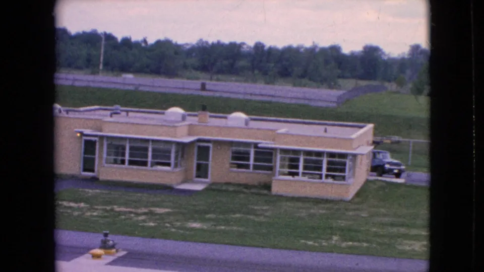 1964 Niagara Falls home-movie frame of the beige building from a slightly shifted handheld angle, with window rows, grassy foreground, road edge, trees, and a dark car at right.