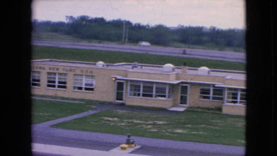 1964 Niagara Falls frame showing the building from another angle, with a central doorway area, rows of windows, a curved walk or drive in the grass, road, and tree line beyond.