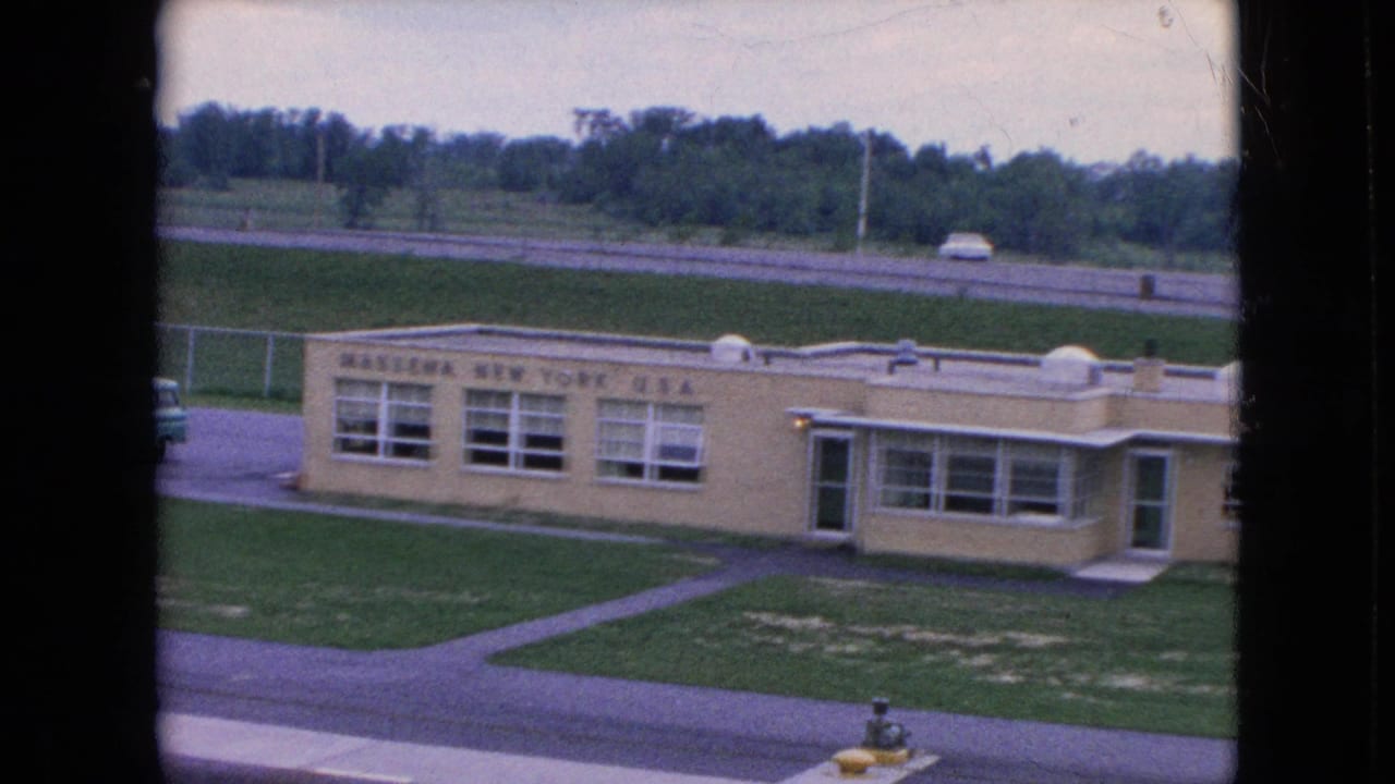 1964 Niagara Falls, New York home-movie frame looking down at a single-story public-looking building with a visible sign, flat roof, lawn, paved paths, and a road behind it.