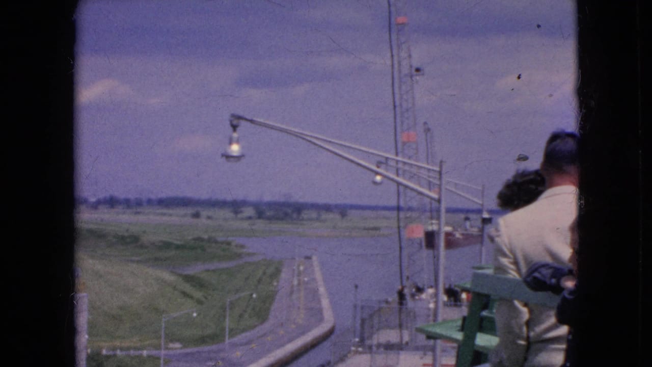 1964 Niagara Falls, New York travel footage frame showing visitors partly visible at an observation area beside railings, with a waterway, curving road, grass, and tall lamp standards below.