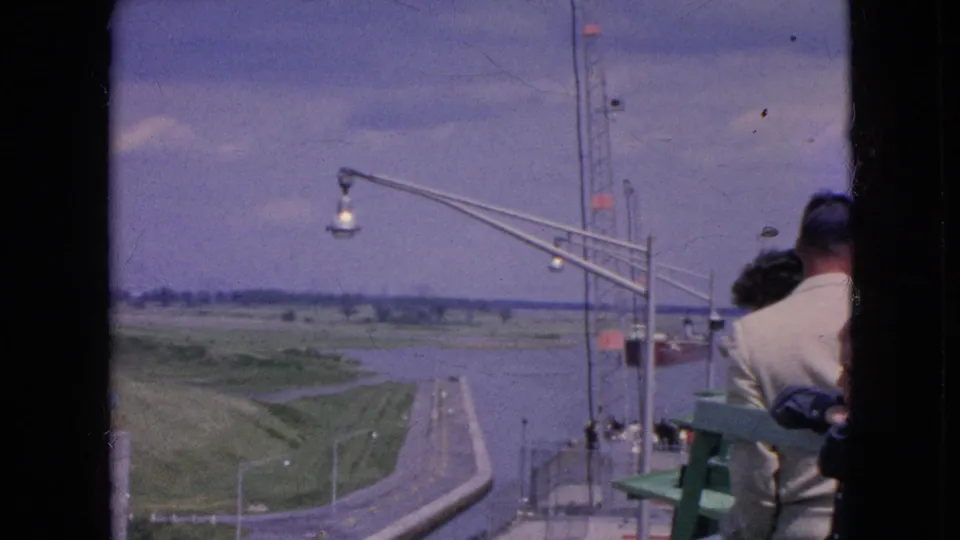 1964 Niagara Falls, New York travel footage frame showing visitors partly visible at an observation area beside railings, with a waterway, curving road, grass, and tall lamp standards below.