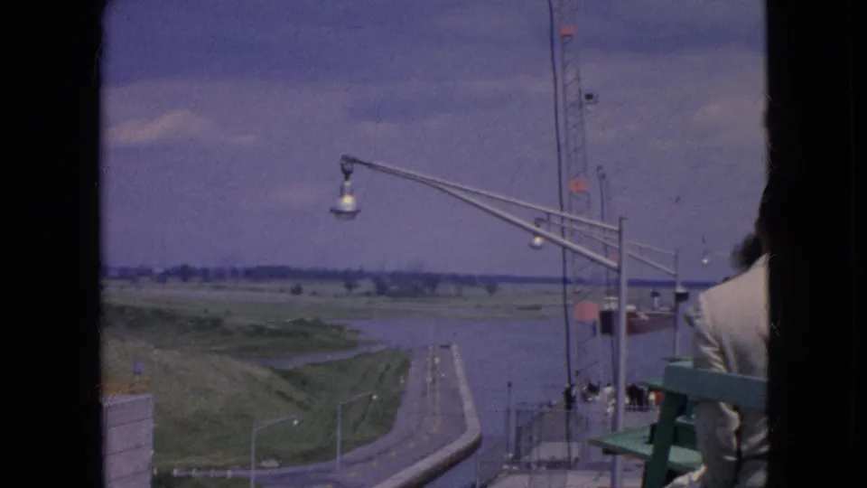 1964 Niagara Falls, New York frame from an elevated visitor vantage point, looking over a canal-side road, grassy embankment, railings, water, and tall street lamps, with a person partially visible at right.