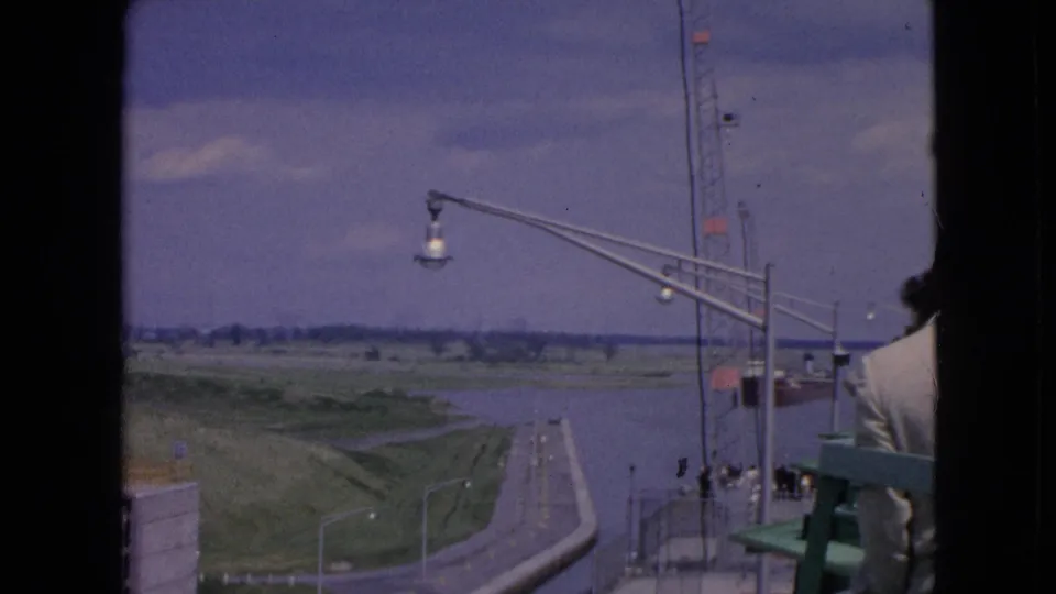 1964 Niagara Falls, New York home-movie frame holding on the elevated overlook, with the waterway to the right, roadway and grass below, tall lamps, railings, and a visitor partly cropped at the frame edge.