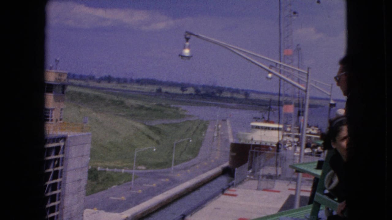 1964 home-movie frame from the United States showing a freighter moving through a concrete lock with a tan service building, waterway, lamps, and partial spectator figures at the edge.