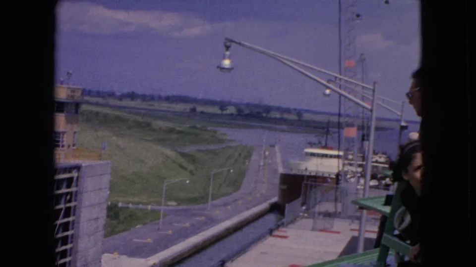 1964 home-movie frame from the United States showing a freighter moving through a concrete lock with a tan service building, waterway, lamps, and partial spectator figures at the edge.