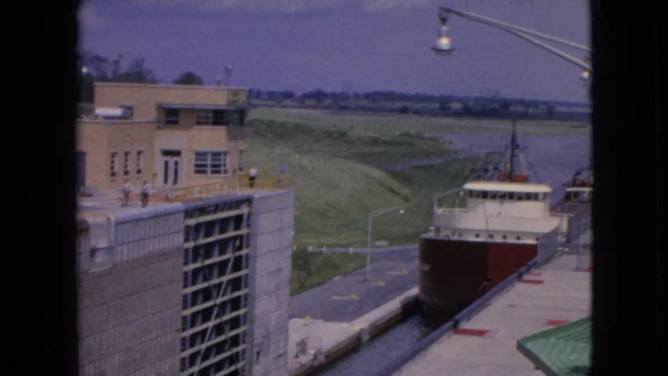 The freighter advances through the lock in 1964 footage, with concrete walls, a tan building, small spectator figures, lamps, a distant waterway, and a green roof edge in view.