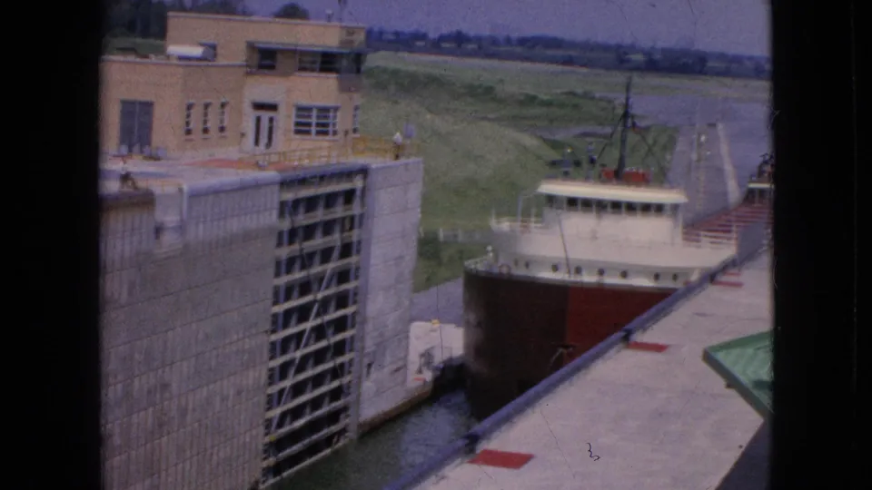 A final 1964 frame shows the freighter continuing through the concrete lock, with the tan building, high lock gates, walkway, railing, grassy embankment, and waterway beyond.