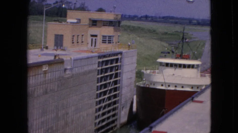 Handheld 8mm view of the freighter progressing through the lock, framed by concrete walls, a control building, and dark film borders.