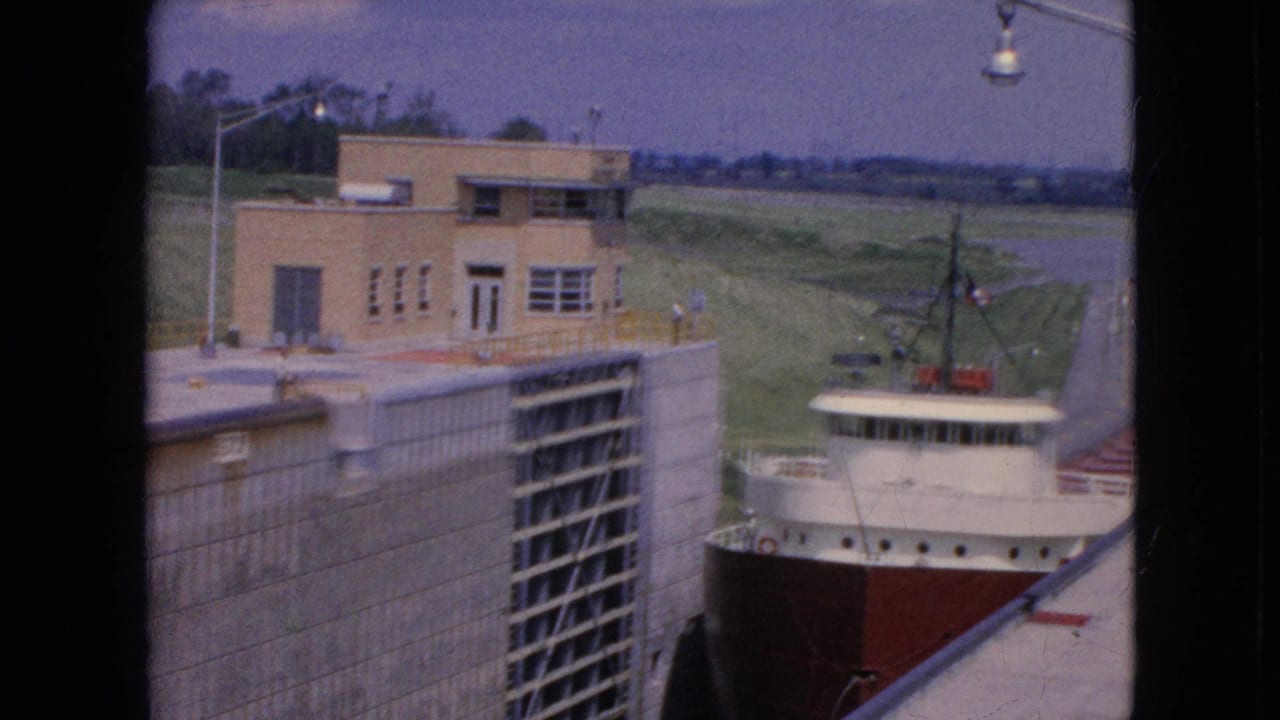 1964 Niagara Falls, New York 8mm frame showing a red-hulled freighter moving through a concrete lock beside a tan control building and grassy embankment.