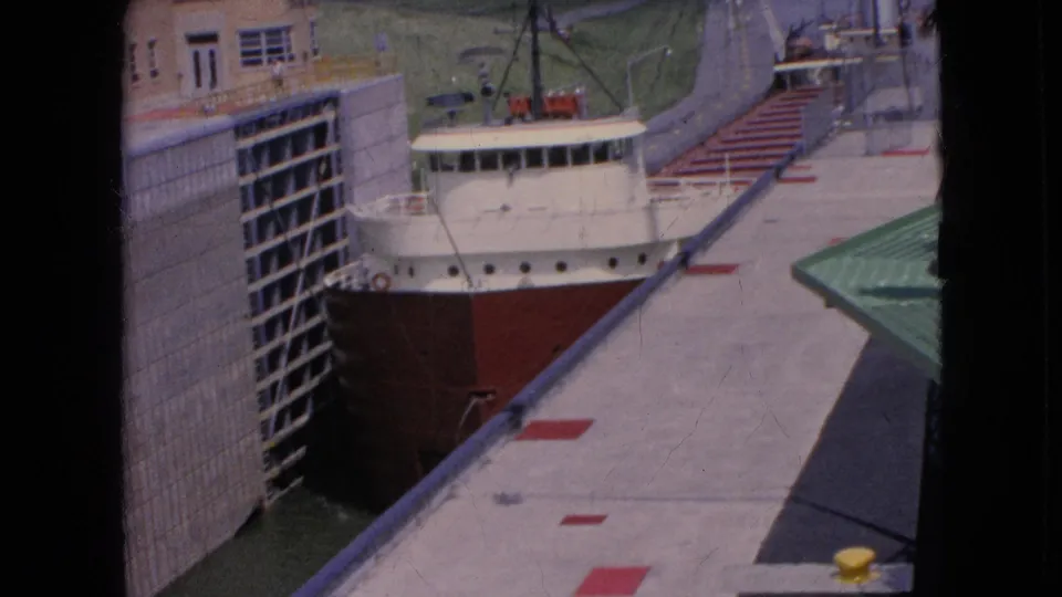 1964 Niagara Falls, New York frame of a freighter moving through the lock beside concrete walls, red deck markings, a green platform, and a small yellow object in the foreground.