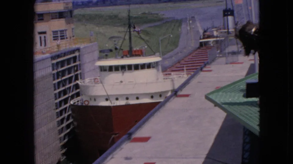 Elevated 1964 home-movie frame of a freighter in a Niagara Falls, New York lock, with concrete walls, dock pavement, and surrounding industrial structures visible.