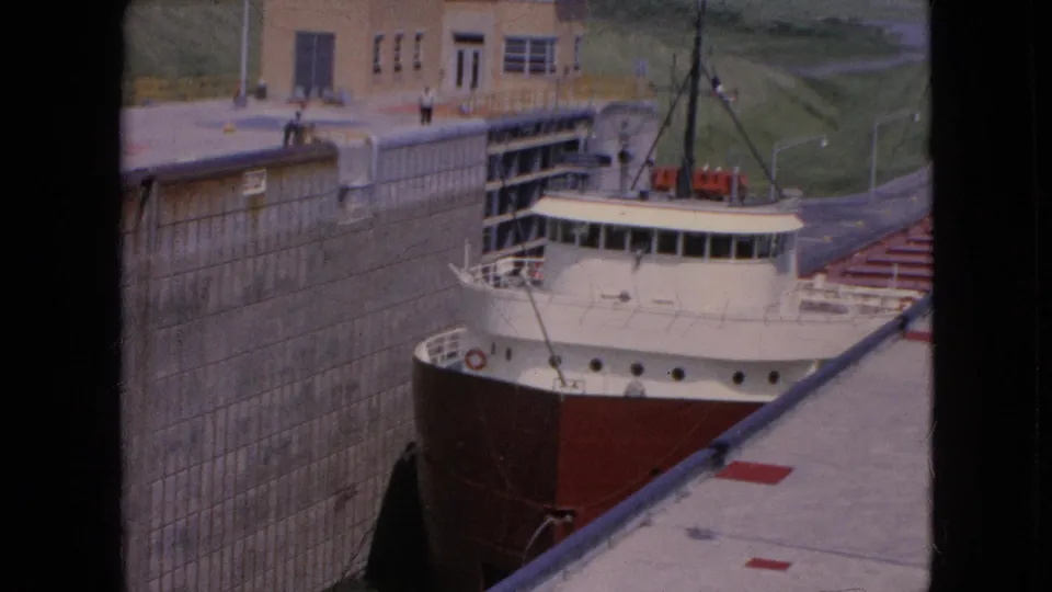 1964 8mm frame from Niagara Falls, New York, showing the freighter close to a high lock wall while small distant people stand on the upper dock area.
