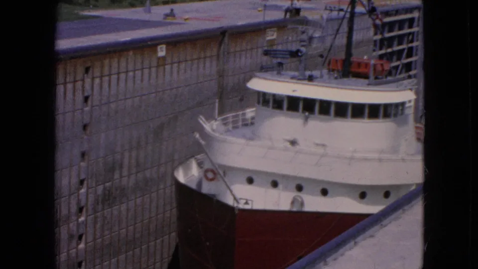 Close 1964 8mm view of the red-and-white freighter moving through the Niagara Falls, New York lock system beside a tall concrete wall and dock edge.