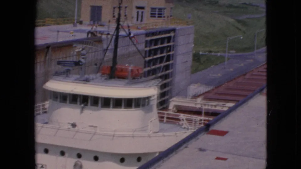 A visitor-shot 8mm view shows the freighter's wheelhouse below lockside buildings, concrete walls, railings, and a roadway or service path beyond the chamber.