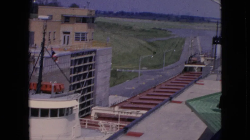 The final frame widens to show part of the freighter below, lockside industrial structures, a paved road, lamps, and grassy terrain around the Niagara Falls lock area.