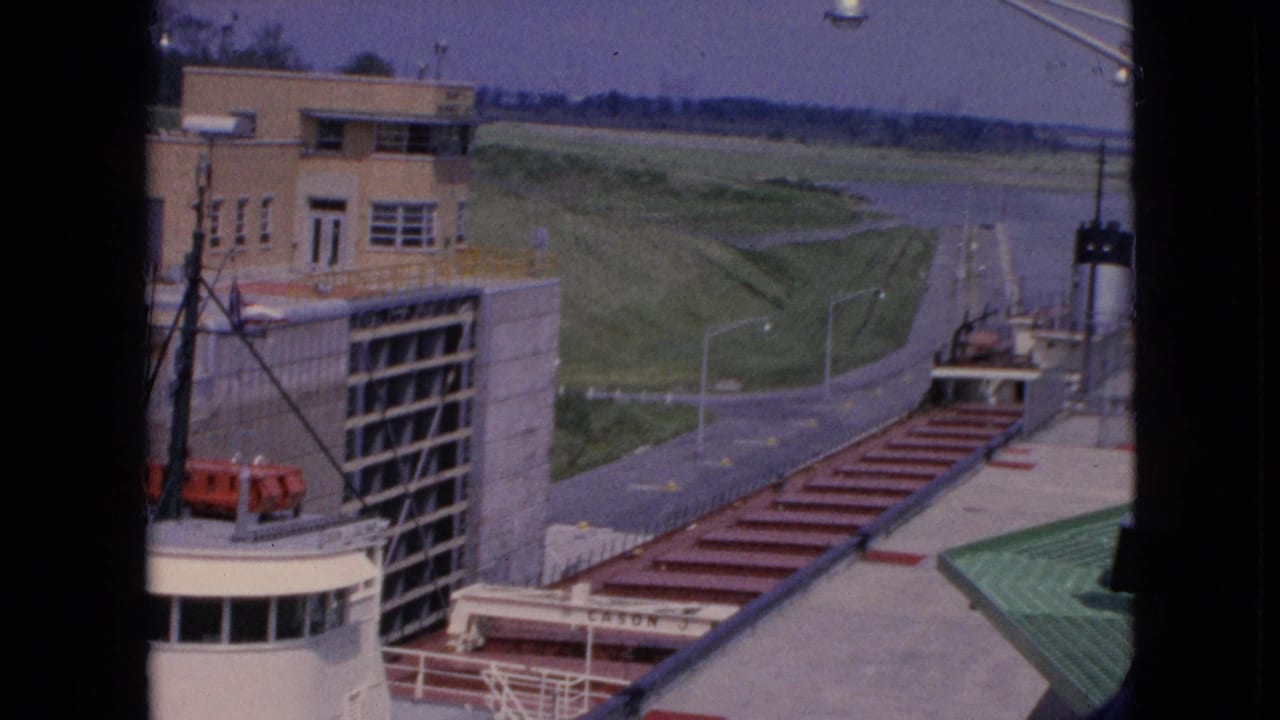 1964 8mm frame from Niagara Falls, New York, showing a white-and-red freighter near a lock wall with dockside structures, a road, and open grass beyond.