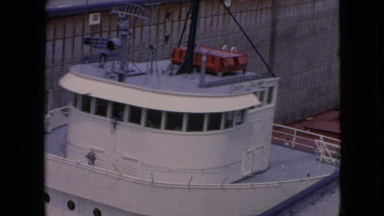 1964 8mm frame of a white freighter's bridge and forward deck inside a concrete lock, with railings, mast rigging, and red deck equipment visible.