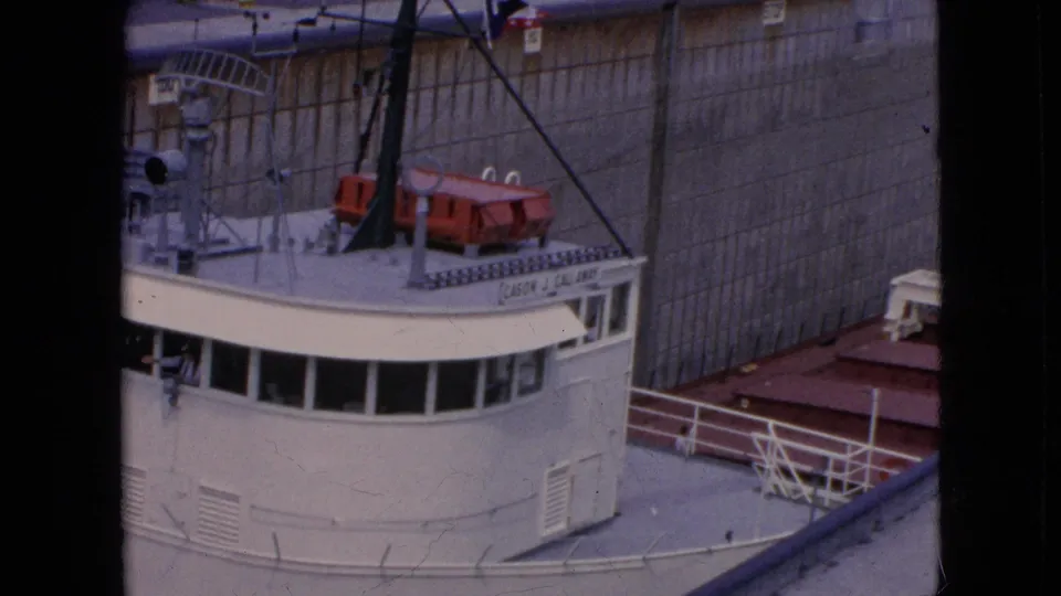 The white freighter's bridge and upper deck appear close to the tall concrete lock wall, with rigging, railings, and red deck equipment visible in the 1964 home-movie frame.