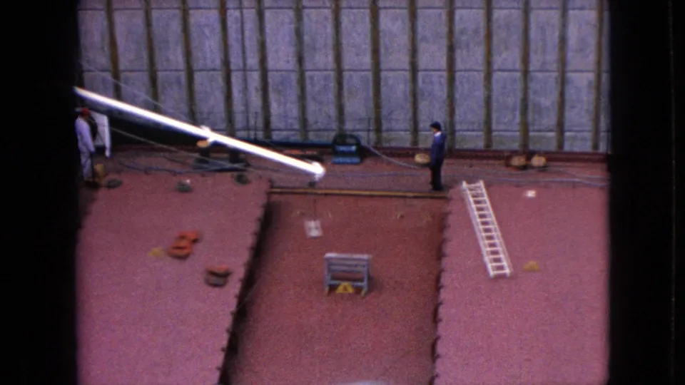 The 1960s home-movie frame shows the red deck of the Cason J. Callaway from above, with a white boom crossing the frame, a distant worker, ladder, and industrial lock wall.