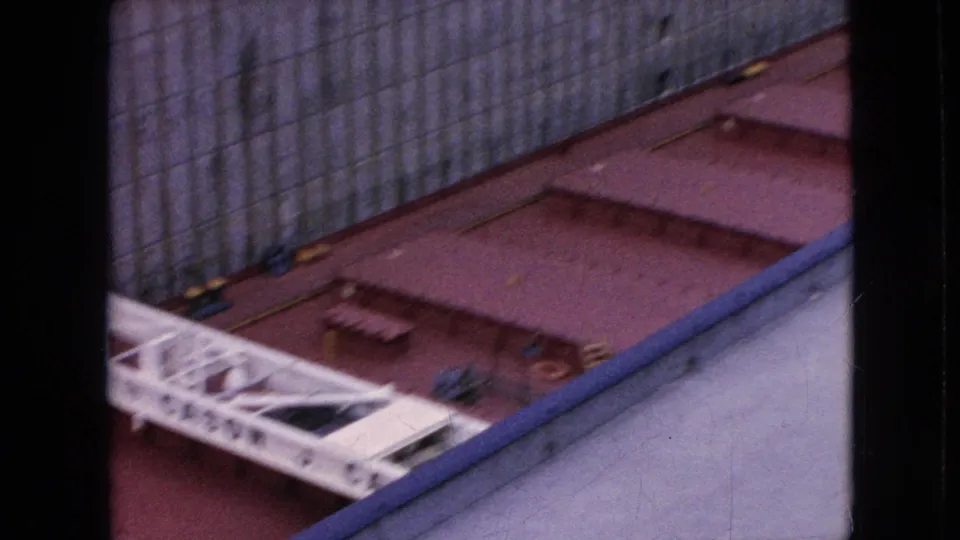 A high-angle 8mm frame shows the vessel deck and tall lock wall from above, with a white structural element, red deck sections, and a blue rail or edge in the foreground.