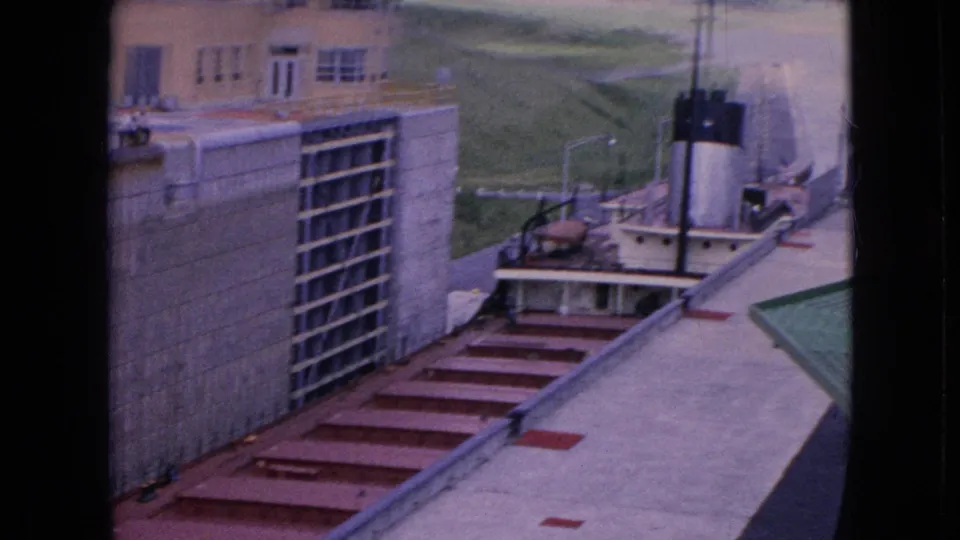 Wider 1964 Niagara Falls power facility view with concrete walls, red metal deck sections, utility equipment, grass beyond the structure, and a small vessel-like form in the distance.