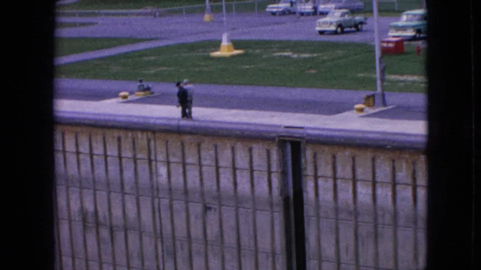 Two distant workers stand on a dock edge above a concrete wall, with roadway, grass, and period cars visible beyond.