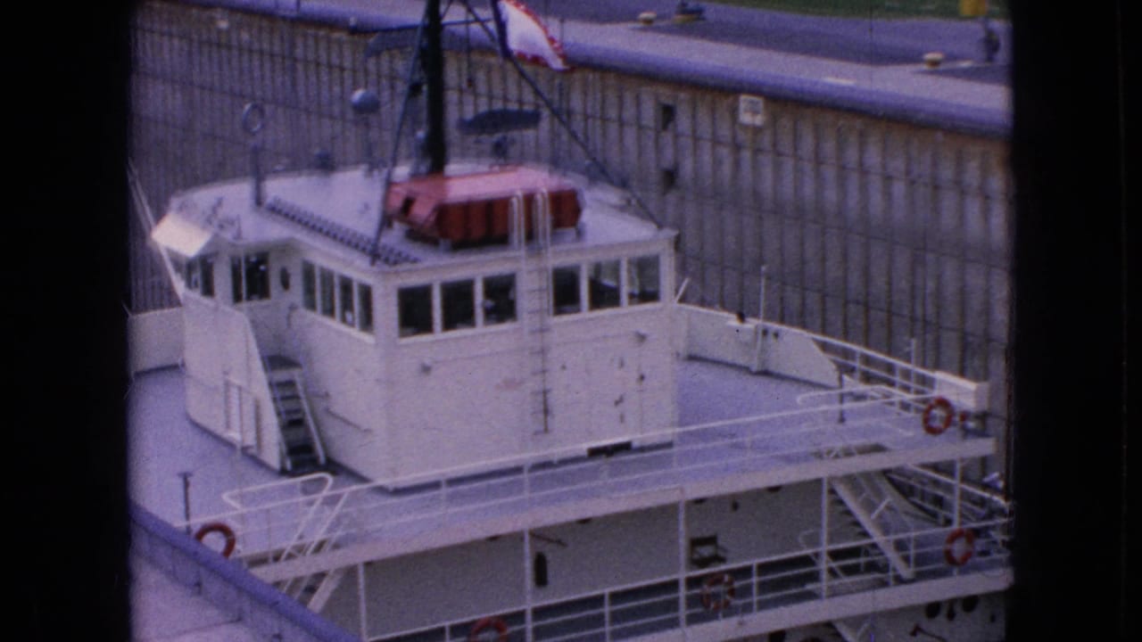 1964 home-movie frame from Niagara Falls, New York, showing a white tugboat with pilothouse windows and red life rings moving beside concrete lock walls.