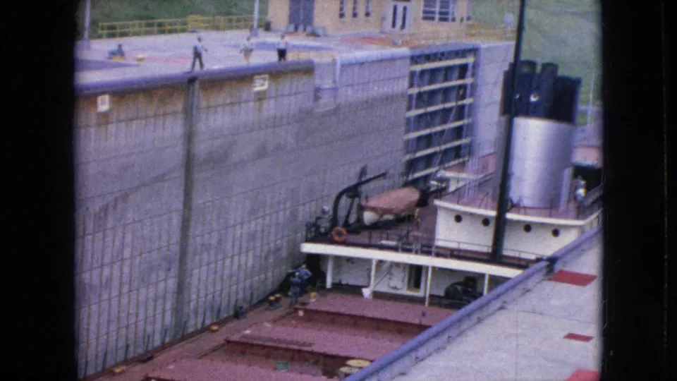 1964 home-movie frame showing the tugboat low in a concrete lock channel while small figures stand along the upper walkway near the lock gate.