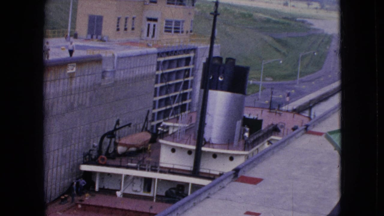 Tugboat in a canal lock with roadway, building, and grassy embankment beyond.
