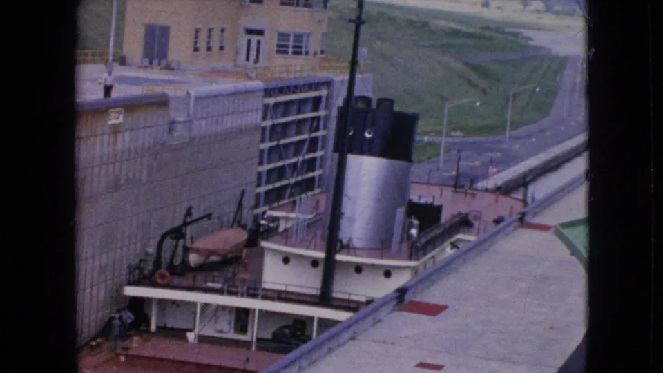 1964 8mm frame of a tugboat with red deck details near a tall lock gate, concrete wall, and service roadway in Niagara Falls, New York.