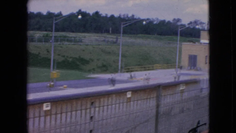 1964 Niagara Falls home-movie frame looking across the top of the lock wall toward a road, grassy embankment, light poles, and distant trees.