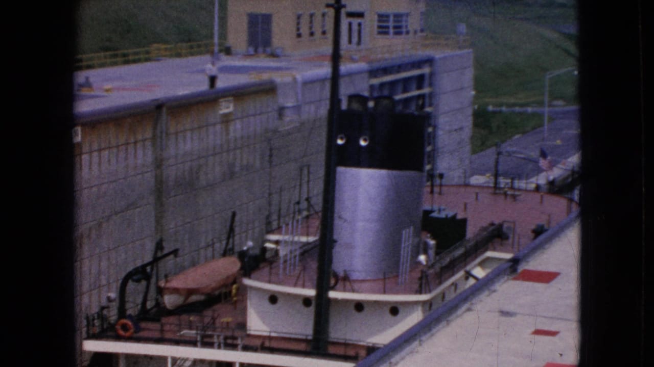 1964 Niagara Falls, New York 8mm frame showing a small boat in a canal lock area with a tall black-and-white smokestack, concrete wall, railings, and industrial building.