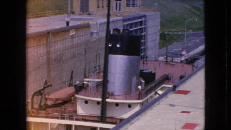 High-angle 1964 8mm view in Niagara Falls, New York showing the boat's smokestack and deck below concrete lock infrastructure, with a road and grassy embankment beyond.