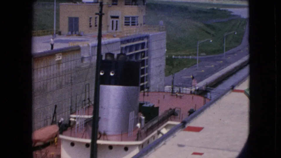 1964 Niagara Falls, New York home-movie frame with the boat's black-and-white smokestack, deck equipment, concrete lock walls, and paved road seen from above.