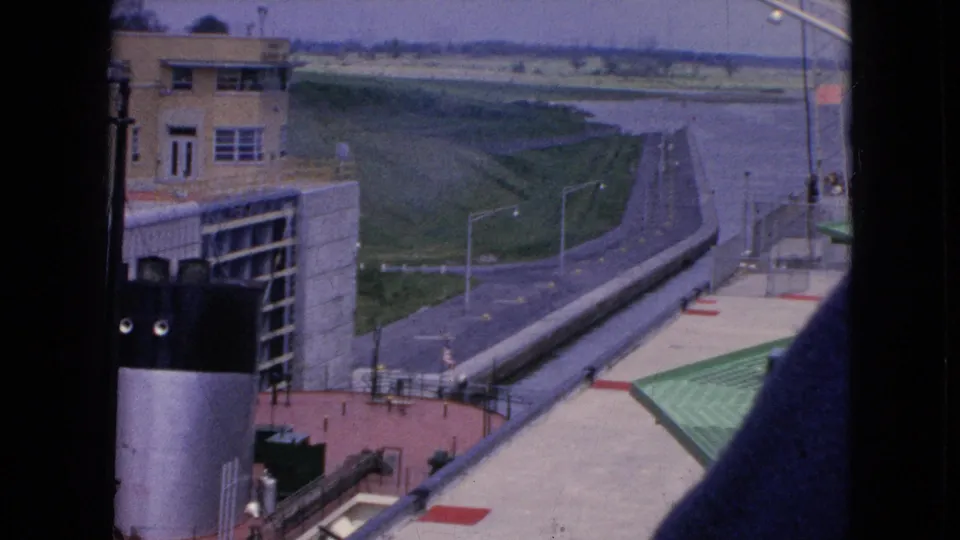 Wider 1964 8mm frame from Niagara Falls, New York showing a boat partly below frame, canal lock walls, a paved access road, grassy slopes, and distant waterway infrastructure.