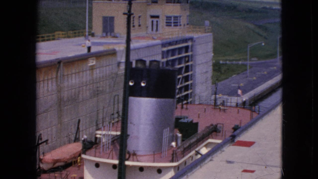 Elevated 1964 home-movie view of a tug or workboat in a canal lock at Niagara Falls, New York, with the lock gate structure, roadway, railings, and grassy slope visible.