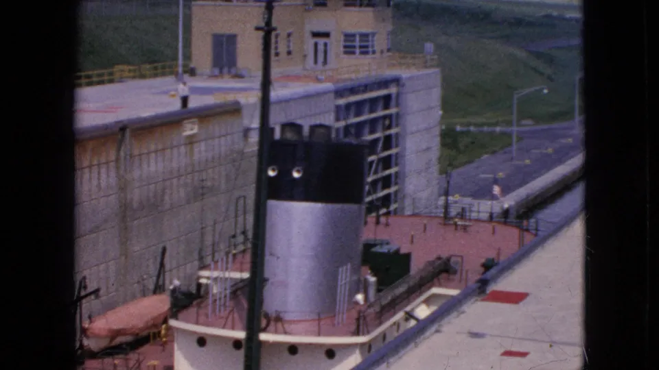 Elevated 1964 home-movie view of a tug or workboat in a canal lock at Niagara Falls, New York, with the lock gate structure, roadway, railings, and grassy slope visible.