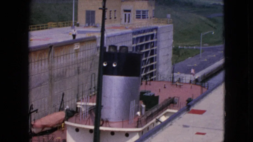 1964 8mm frame showing the tug or workboat closer in the lock, with the black-and-silver smokestack, concrete wall, deck railings, and canal-side roadway visible.