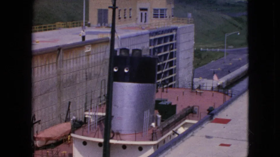 Final 1964 frame from the Niagara Falls canal lock view, showing the tug or workboat aligned with the lock wall, a control building, railings, roadway, and a small American flag.