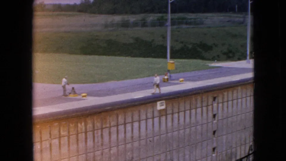 Distant pedestrians walk along the lock walkway above a concrete wall and grassy bank in 1964 Niagara Falls, New York home-movie footage.