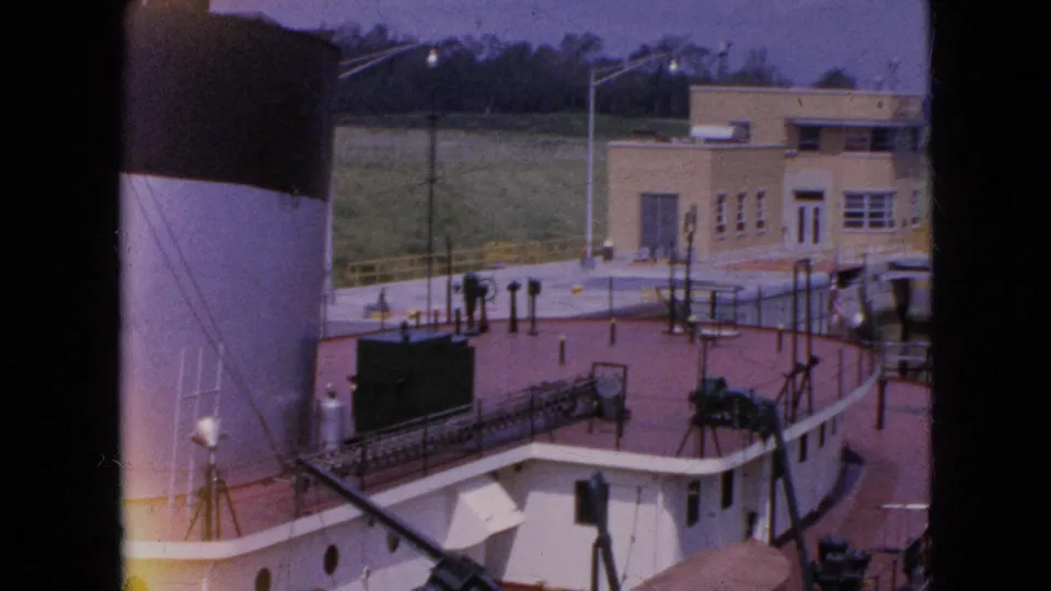 The camera looks across the vessel deck toward the lockside control building, showing railings, fittings, poles, and industrial deck structures in faded 8mm color.