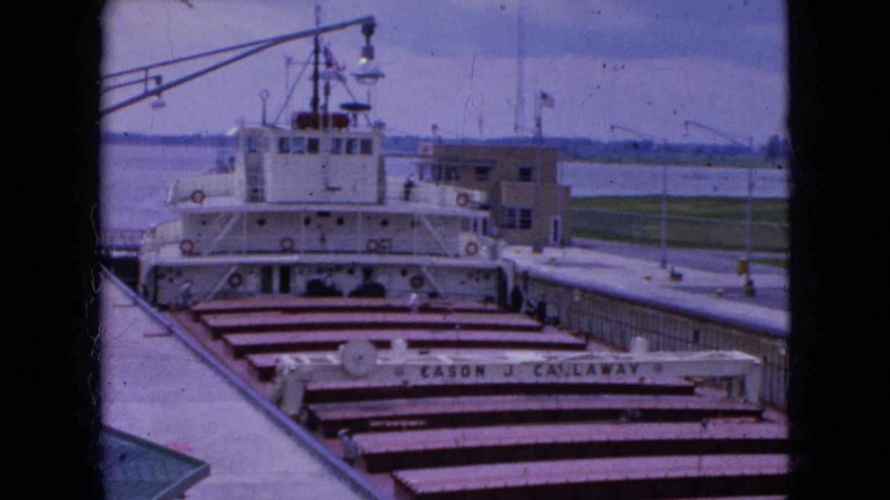 1964 8mm frame from Niagara Falls, New York showing a large white freighter in a canal lock, with cargo hatch covers, railings, water, and a dockside building in view.