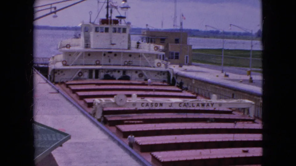 1964 8mm home-movie frame showing a white freighter in the Niagara Falls, New York lock area, with hatch covers, railings, water, and dock structures visible.