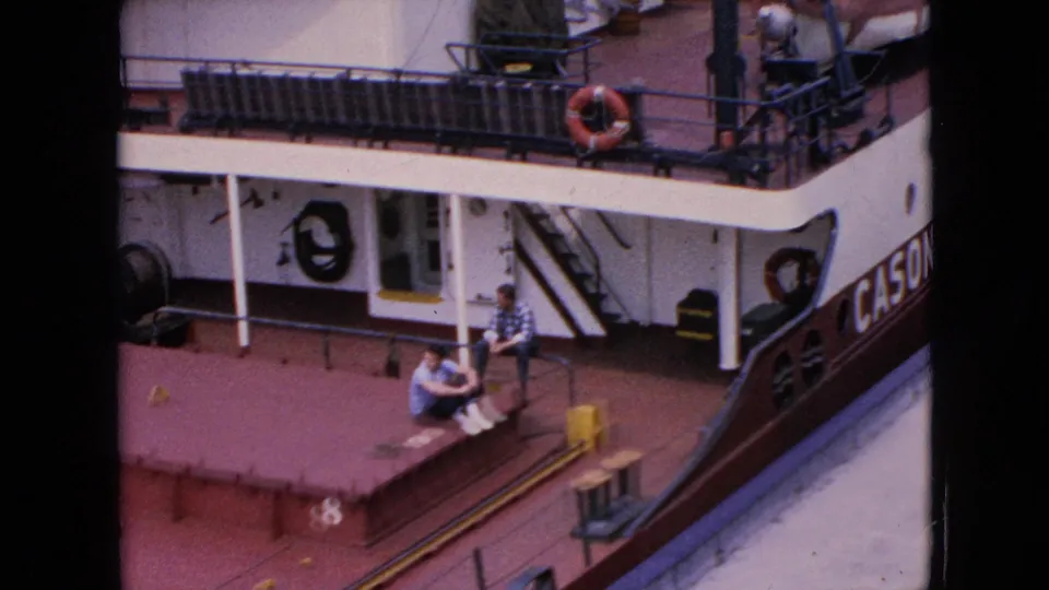 1964 Niagara Falls home-movie frame looking down at two men seated near the docked Cason J. Callaway riverboat, with deck railings and partial vessel lettering visible.