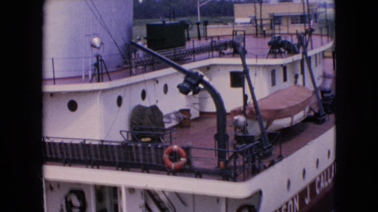 1964 8mm frame showing a close dockside view of the freighter deck, with railings, a covered boat, life preserver, white superstructure, and harbor equipment visible.