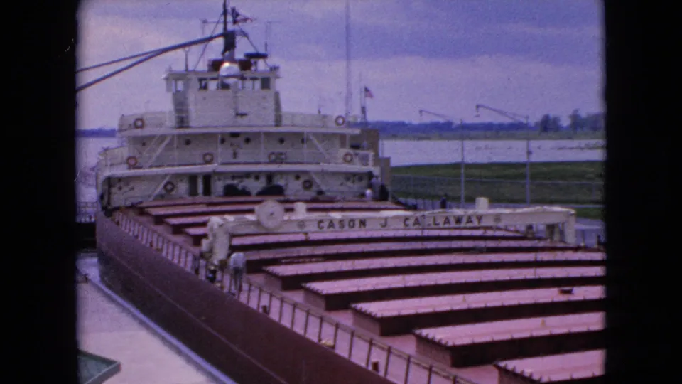 1964 8mm frame showing the Cason J. Callaway from a slightly front-facing dockside angle, with cargo hatches, bridge structure, railings, and shoreline beyond the water.